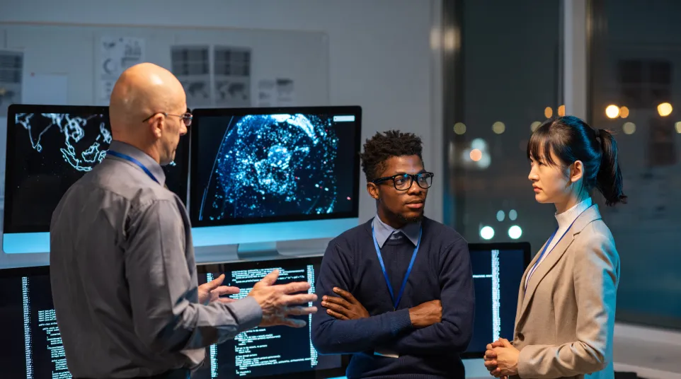 Three professionals discussing data analytics in a modern office environment with multiple monitors.