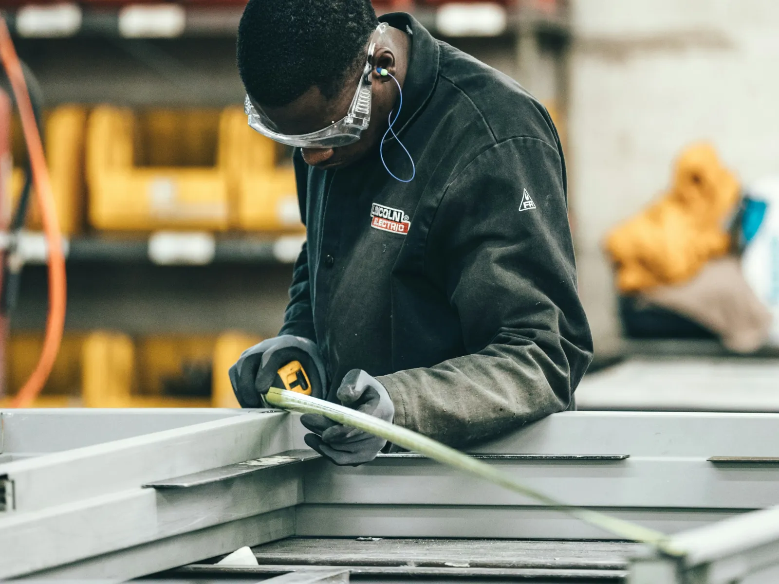 Worker wearing safety glasses and gloves measuring metal frame using a tape measure in industrial workshop.
